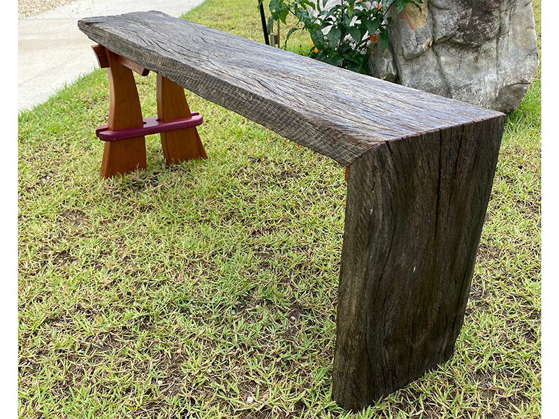 Weathered mahogany waterfall bench with sculpted split leg and purple heart accents. Natural setting.